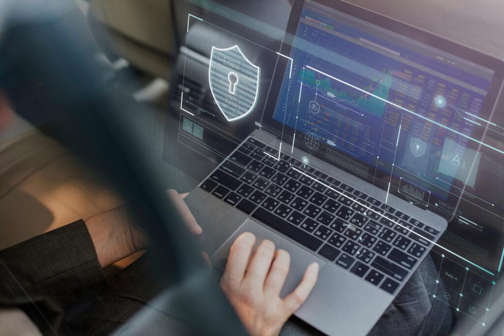 A person working on a laptop with translucent digital security graphics overlaid on the screen, including a shield with a keyhole and various data charts, giving a high-tech cybersecurity atmosphere.