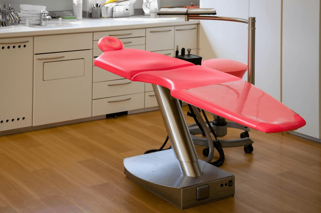 A sleek, modern medical examination room featuring a bright red, wing-shaped exam chair with a metallic base, positioned on polished wooden flooring. Behind it are clean white cabinets, drawers, and a countertop lined with neatly organized dental or medical tools and supplies. Soft, even lighting and uncluttered surfaces give the space a sterile, professional atmosphere.