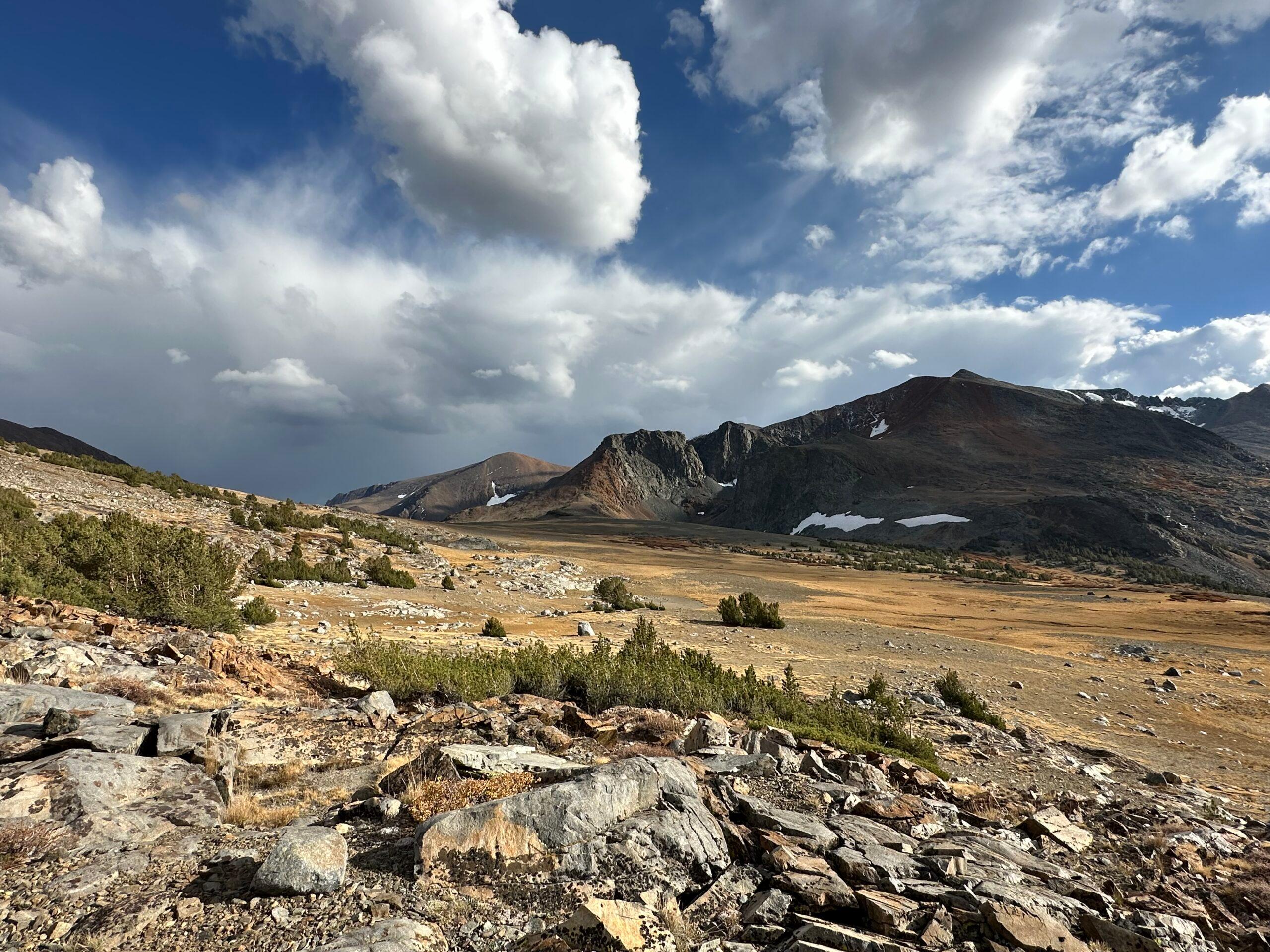 Vast rocky mountain valley under a dramatic sky filled with large billowing clouds, with patches of snow on distant ridges and scattered shrubs across the sunlit terrain.