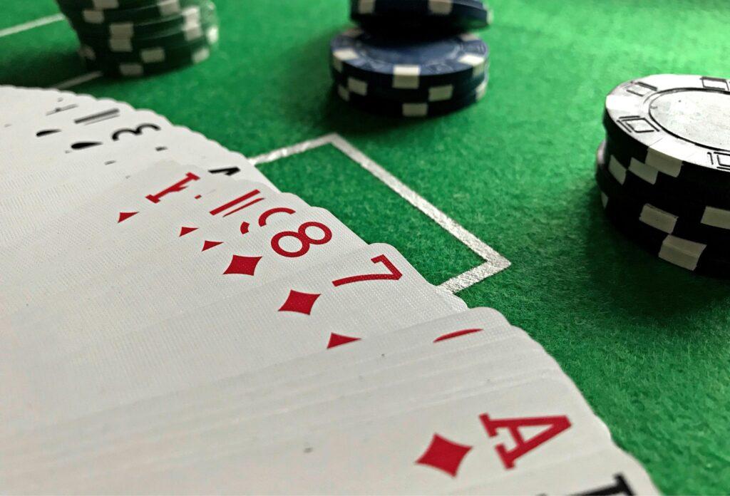 Close-up of playing cards spread out on a green poker table, with red diamond suits visible and stacks of blue, white, and black poker chips in the background.