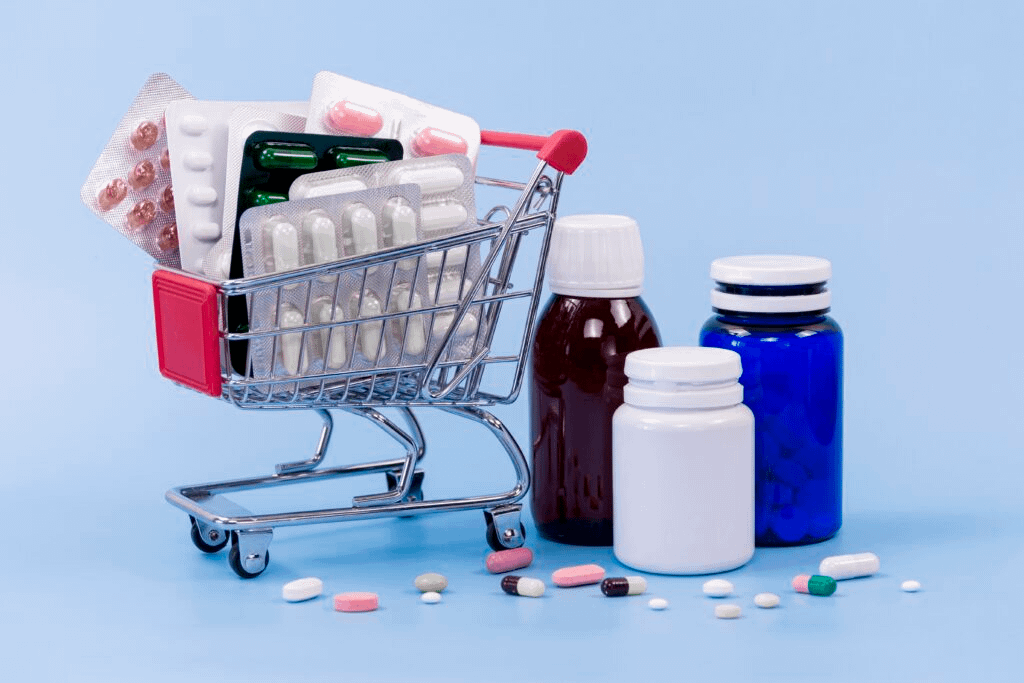 A small metal shopping cart filled with various blister packs of pills, positioned beside several medicine bottles on a light blue surface, with loose tablets and capsules scattered in the foreground.