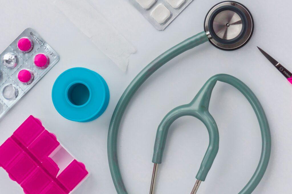A flat lay of medical items including a teal stethoscope, blister packs of white and bright pink pills, a pink pill organizer, a roll of medical tape, and small scissors arranged on a light surface.