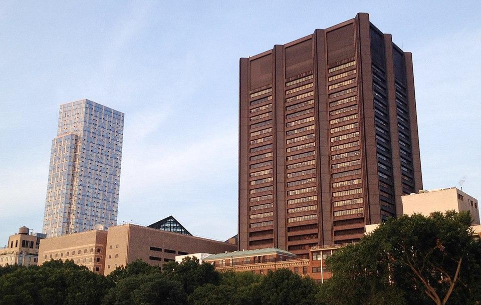 A cluster of tall buildings against a clear sky, including a modern glass high-rise and a large, dark, rectangular tower, with trees in the foreground.