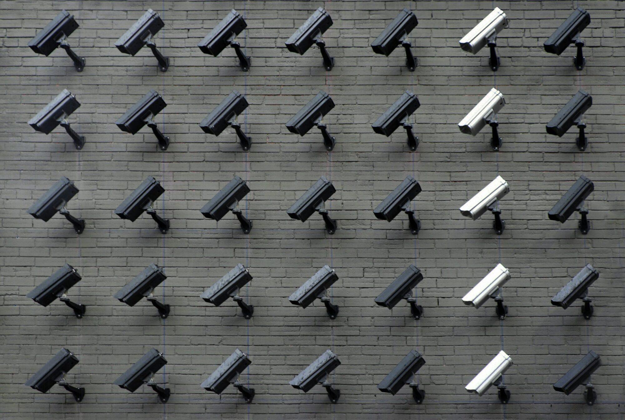 Multiple security cameras mounted in a grid pattern on a gray brick wall, all angled in the same direction with alternating black and white casings.