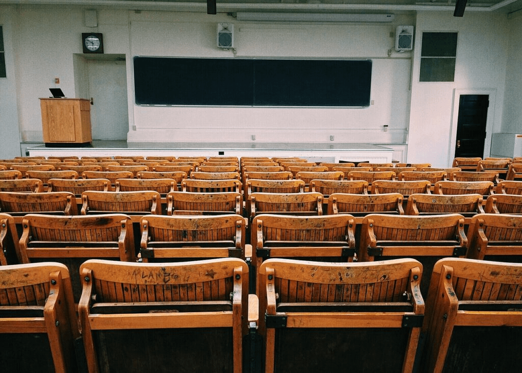 An empty lecture hall filled with rows of worn wooden seats facing a large chalkboard and a podium at the front.