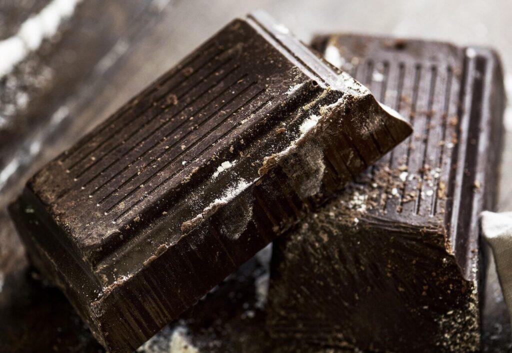 Close-up of rough, partially broken blocks of dark chocolate, coated with a light dusting of cocoa powder and whitish bloom, showing textured ridges and small crumbs scattered around.