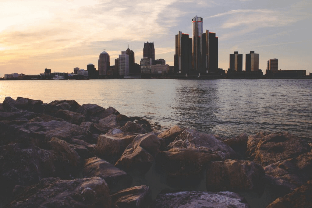 Rocky shoreline at sunset with calm water in the foreground and a distant city skyline silhouetted against a softly colored sky.