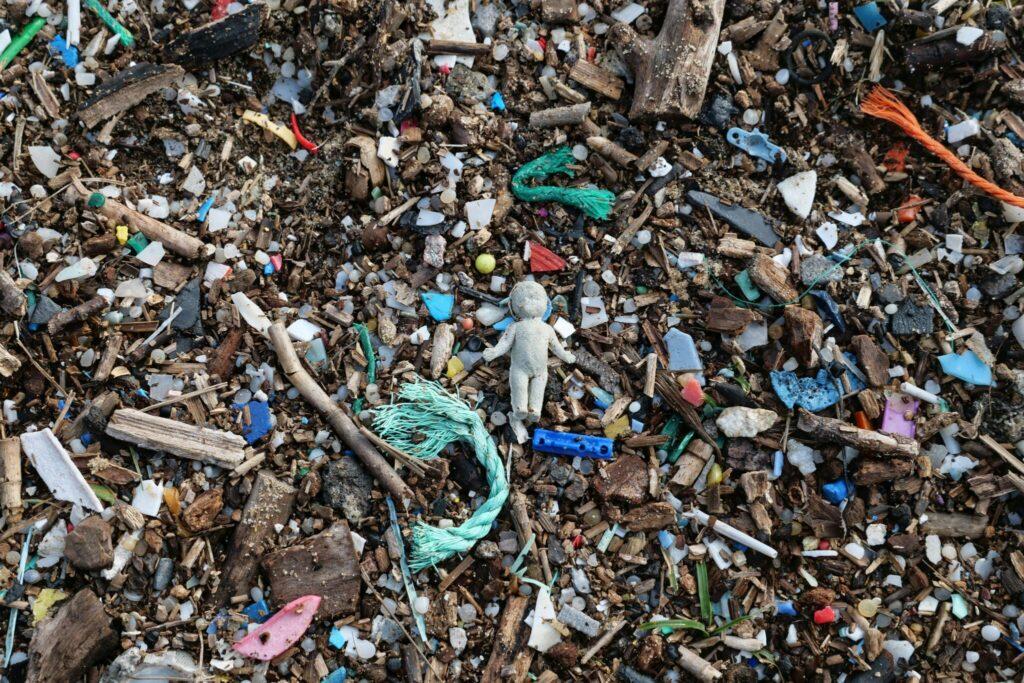 Discarded debris and microplastics scattered across the ground, with a small weathered doll lying among pieces of wood, rope, and colorful plastic fragments.