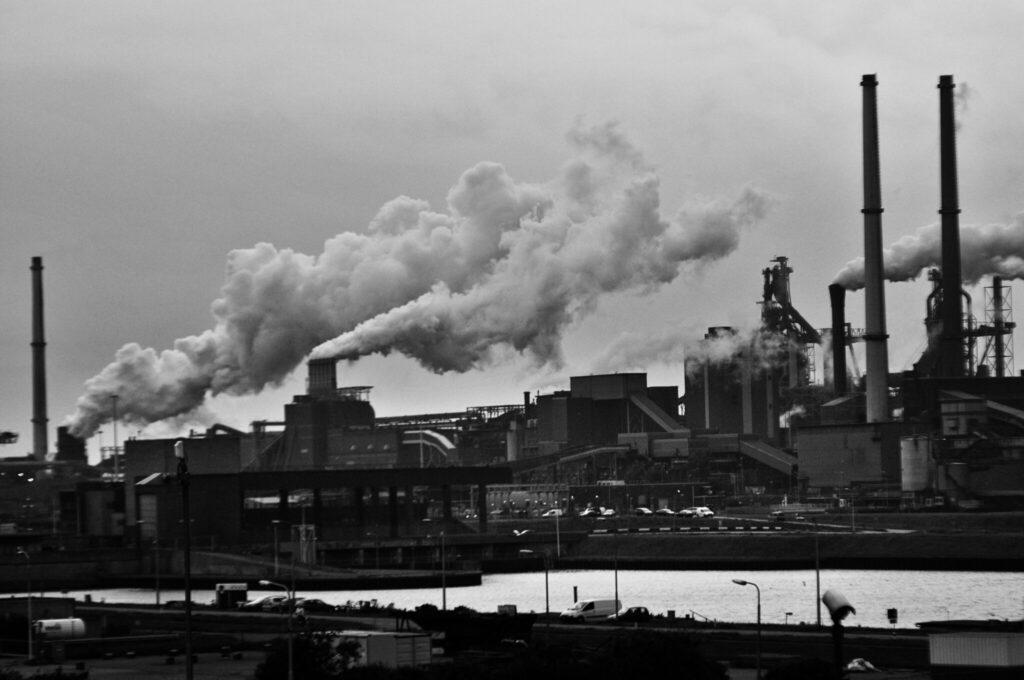 Black and white image of an industrial complex with multiple smokestacks releasing thick plumes of smoke into the sky above a riverfront area.