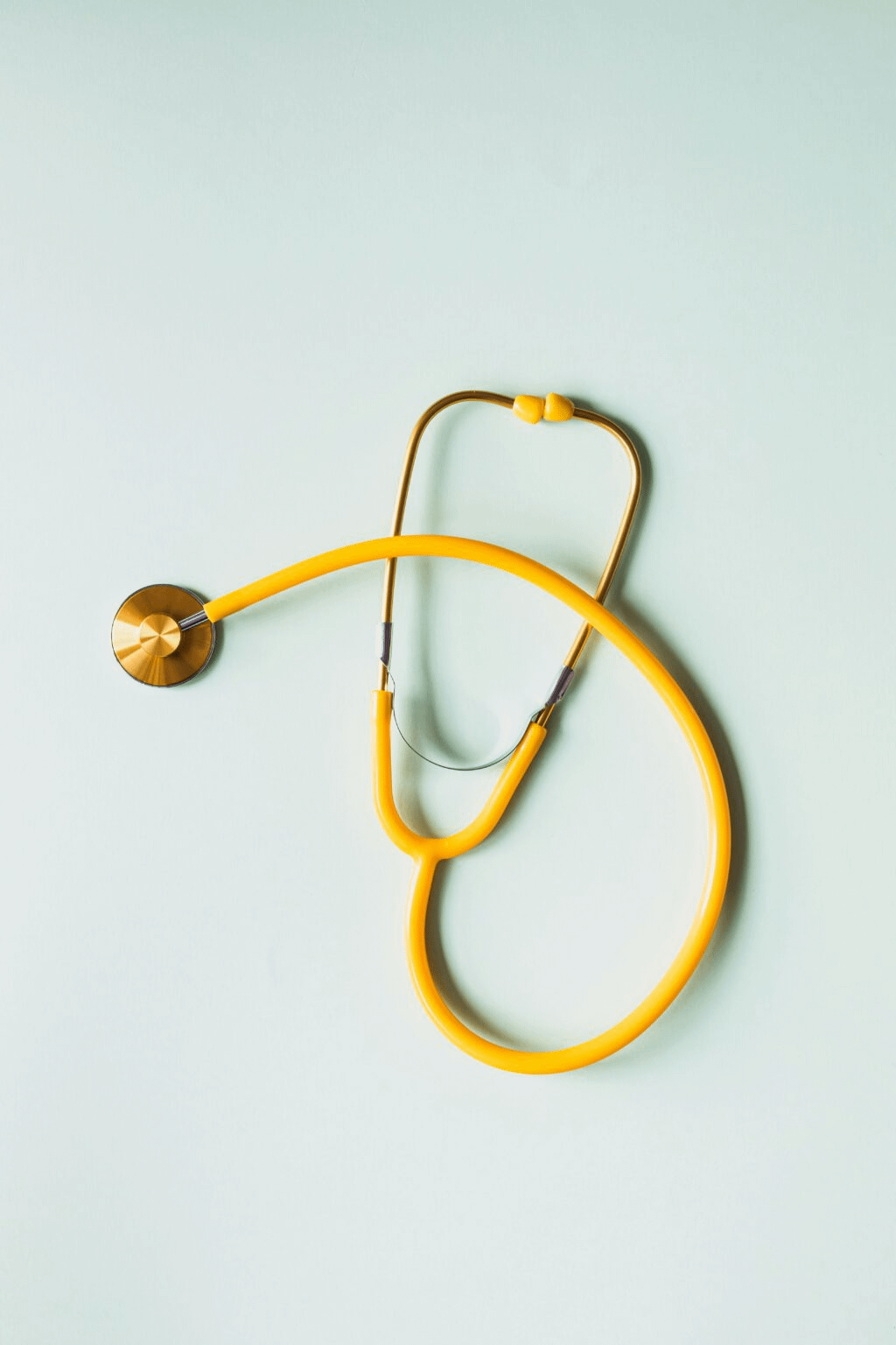 Bright yellow stethoscope with metal chest piece lying on a light pastel surface, the tubing loosely curved and the earpieces resting at the top.