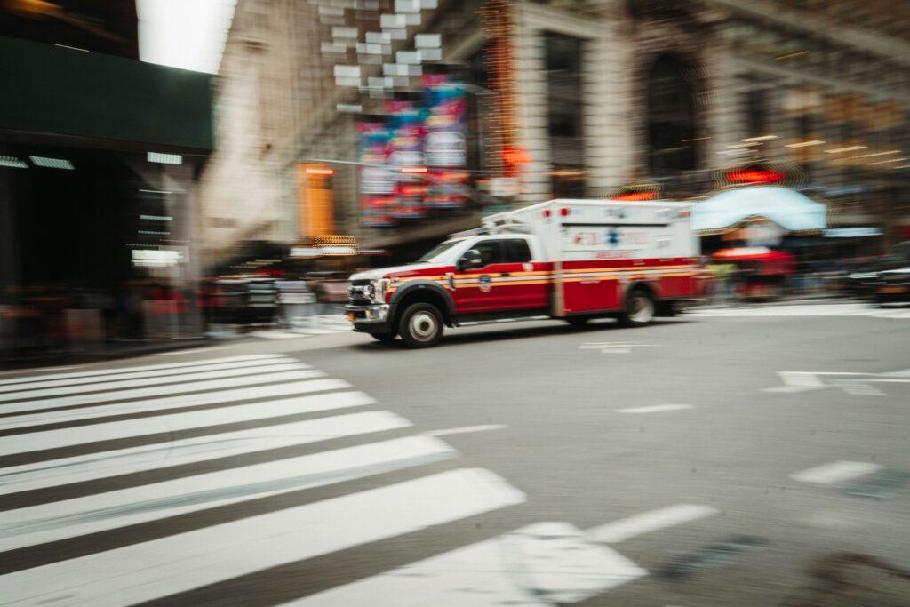 A red and white ambulance speeding through a busy city intersection, captured with motion blur as it passes crosswalk lines and blurred buildings.